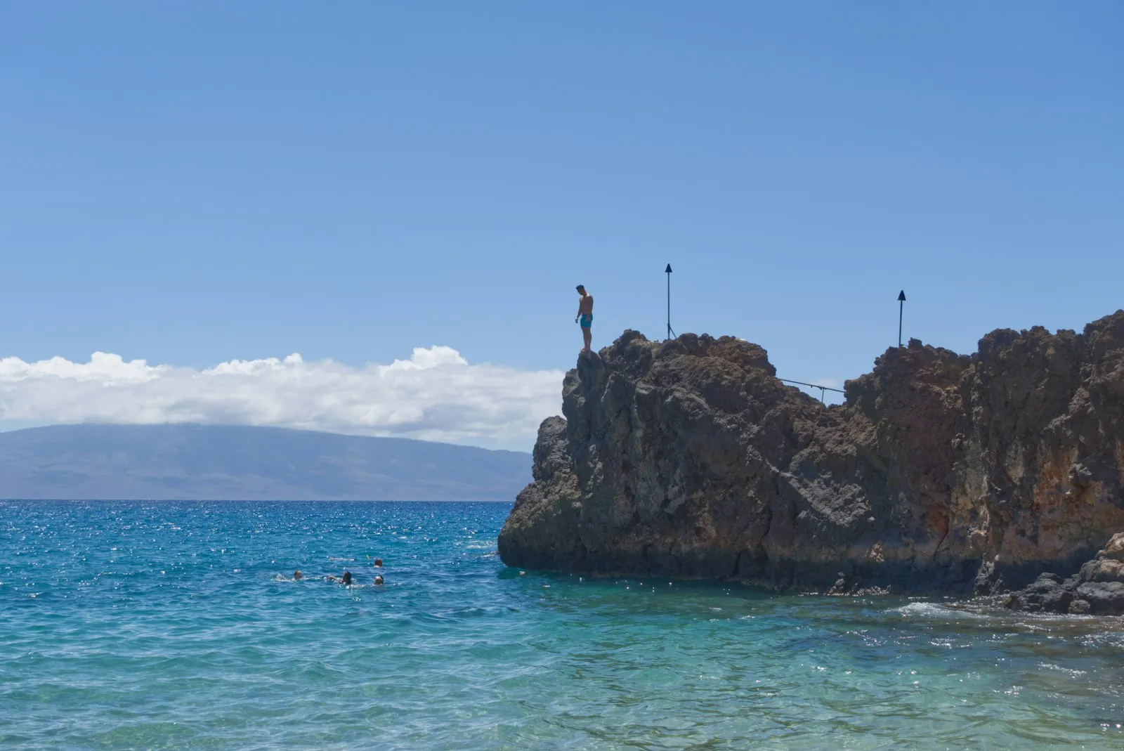 Kāʻanapali 北端的 Black Rock 黑色熔岩岬角，一人站在崖顶，浅绿海水中有小小的游泳者，背景是 Lānaʻi