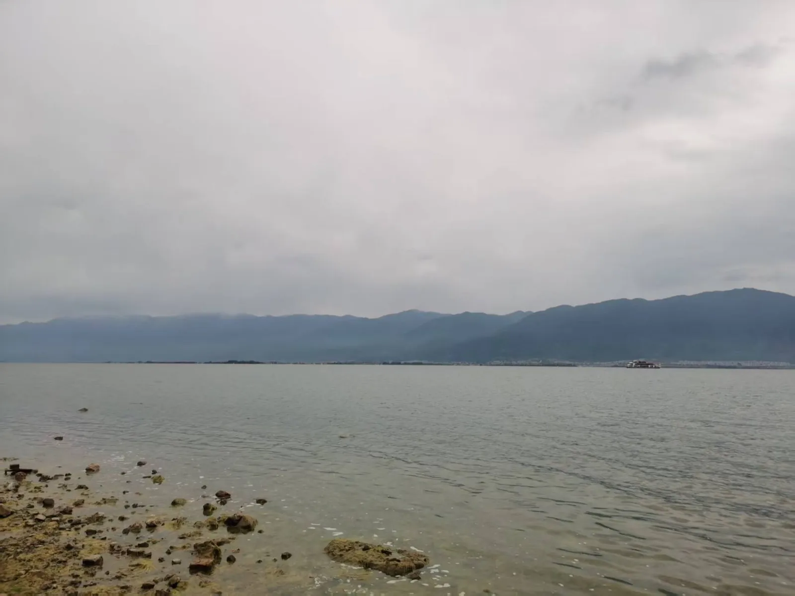 阴天的洱海，水面平静，远处群山隐在云雾中，近岸是布满礁石的浅滩