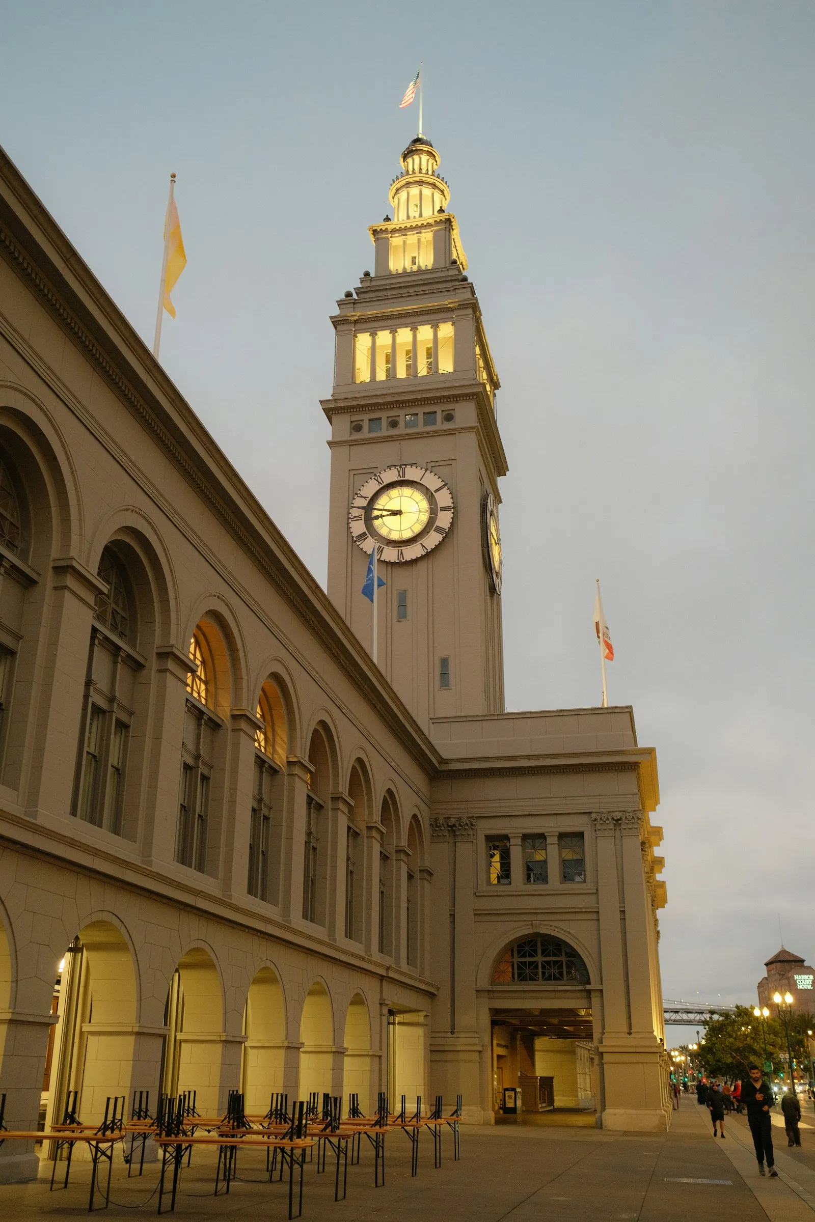 黄昏亮灯的 Ferry Building 钟楼与拱廊立面，远处是海湾大桥