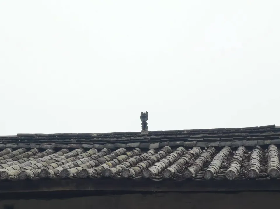束河古镇一处灰瓦屋顶，屋脊正中蹲着一只造型古朴的瓦猫，背景是空旷的天空