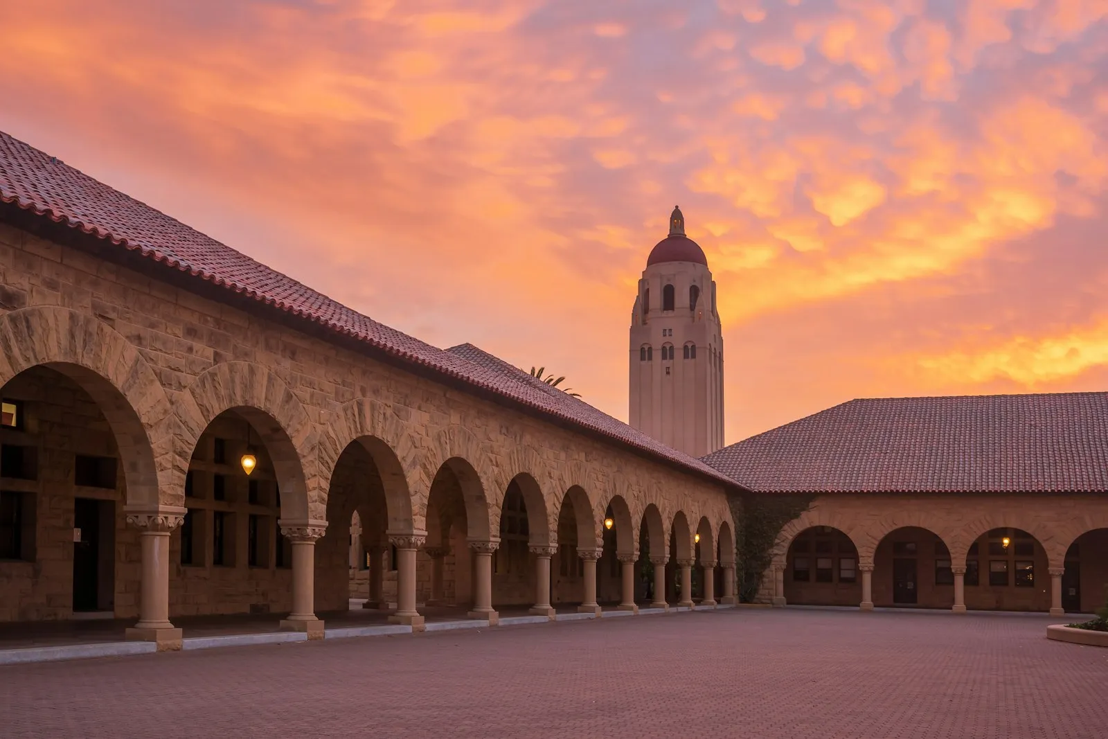 斯坦福主方庭的砂岩拱廊与红瓦屋顶，Hoover Tower 立在粉橙色的晚霞里
