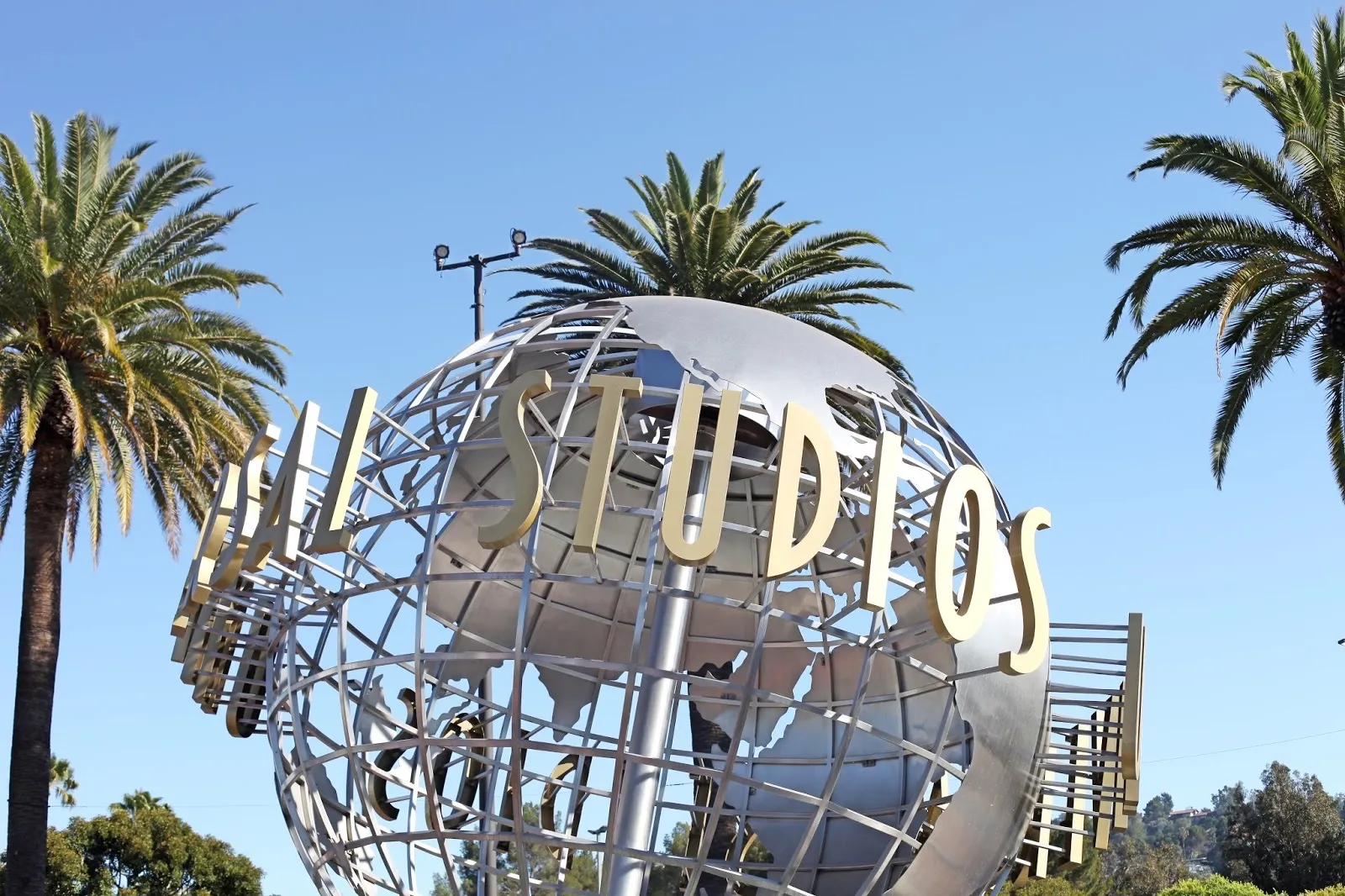 Universal Studios Hollywood 门口的银色地球仪雕塑,金色 Universal Studios 字样,背景是棕榈树