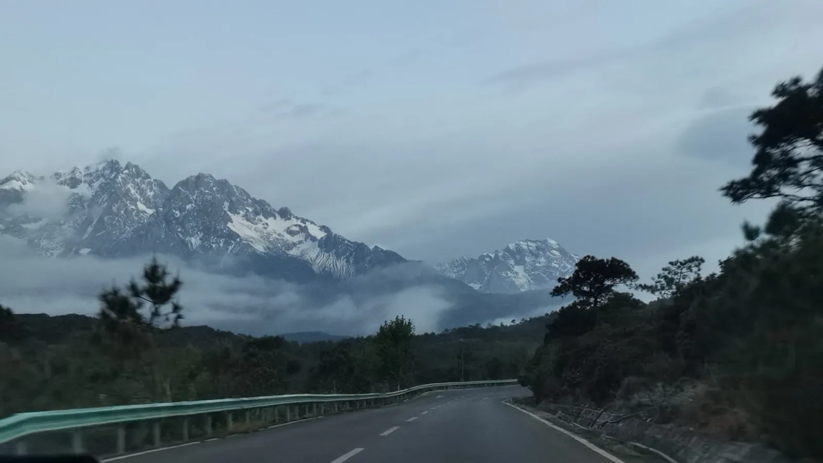 清晨公路尽头的玉龙雪山，雪峰被云雾半掩，山下是松林与蜿蜒山路