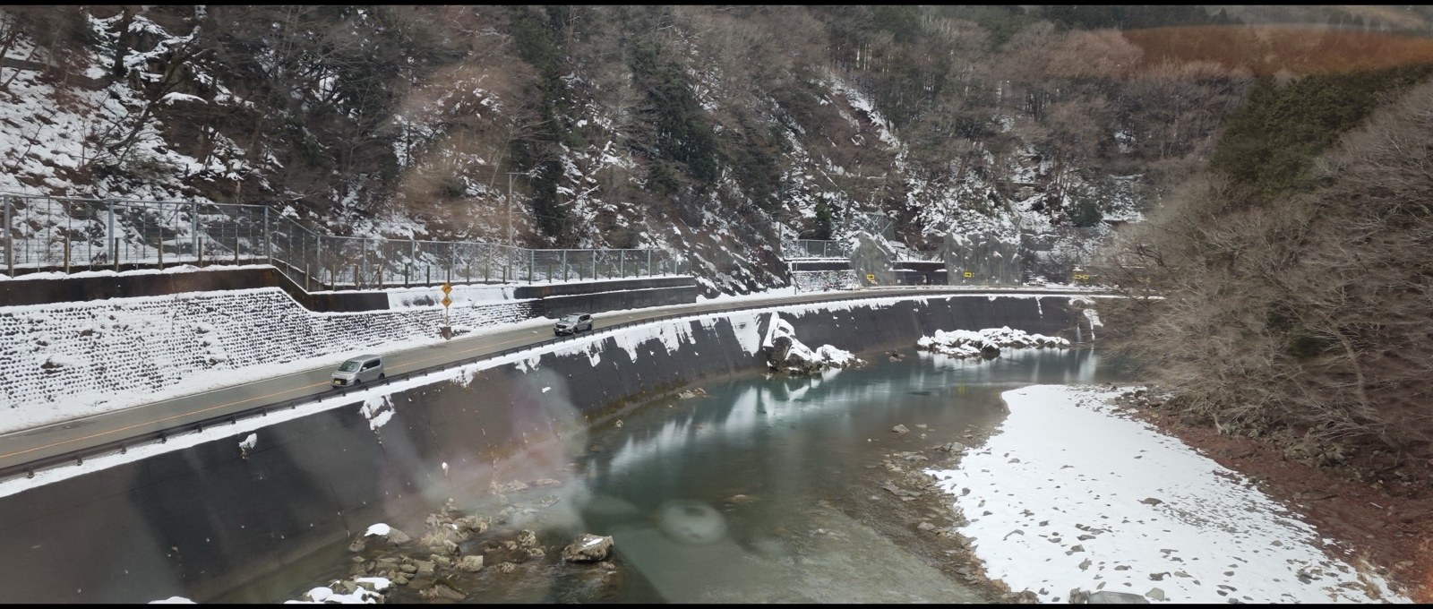 飛驒号列车窗外，一条河流蜿蜒穿过雪谷，沿河公路与对岸山坡都覆着积雪