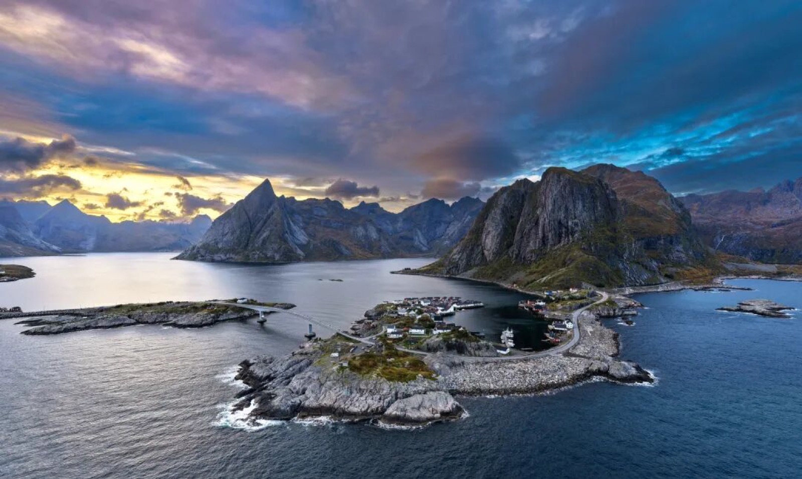 罗弗敦雷纳峡湾的航拍全景，尖耸的山峰环抱海湾，红顶渔村点缀在礁石半岛上，远处天际是橙紫色的霞光
