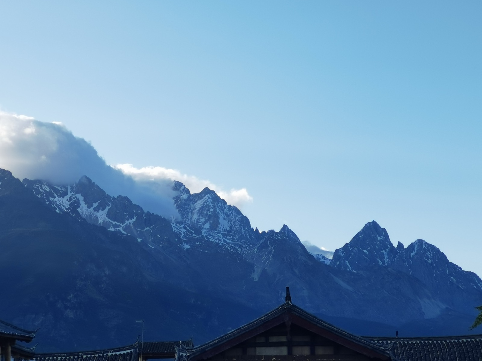 白沙古镇的青瓦屋顶之上，玉龙雪山的雪峰在蓝天下清晰可见，山顶绕着一缕白云