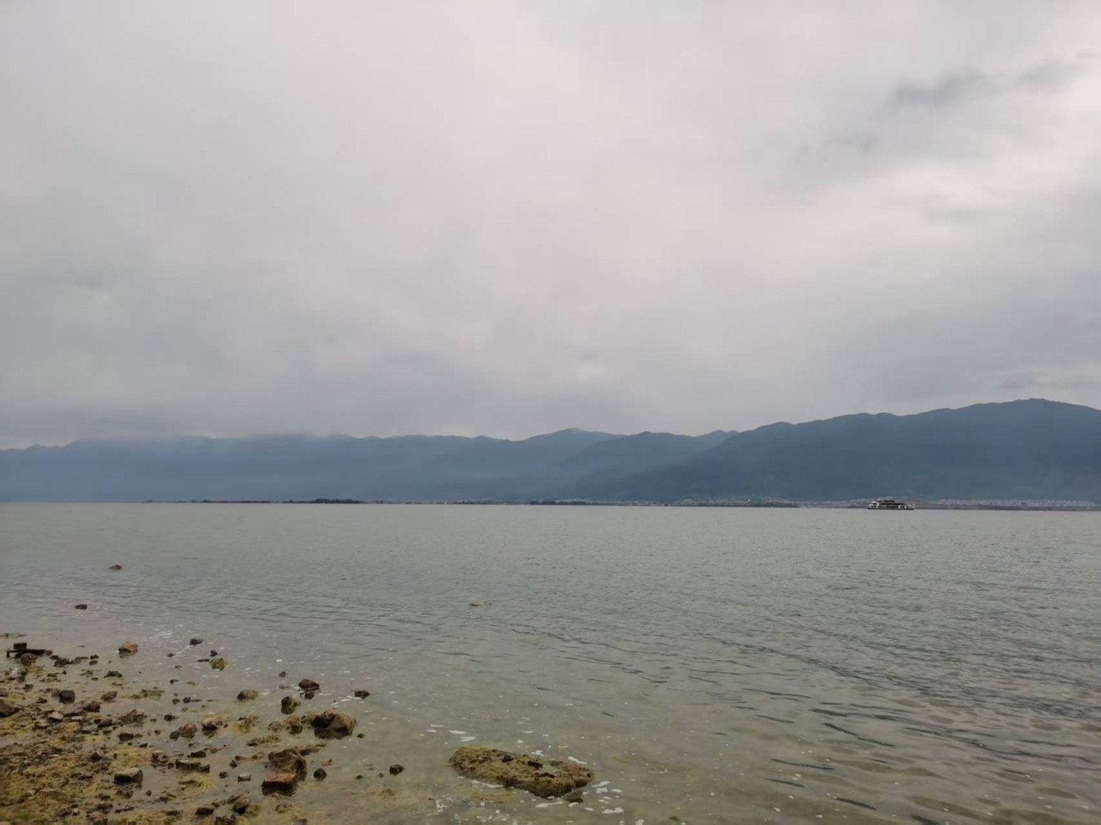 阴天的洱海，水面平静，远处群山隐在云雾中，近岸是布满礁石的浅滩