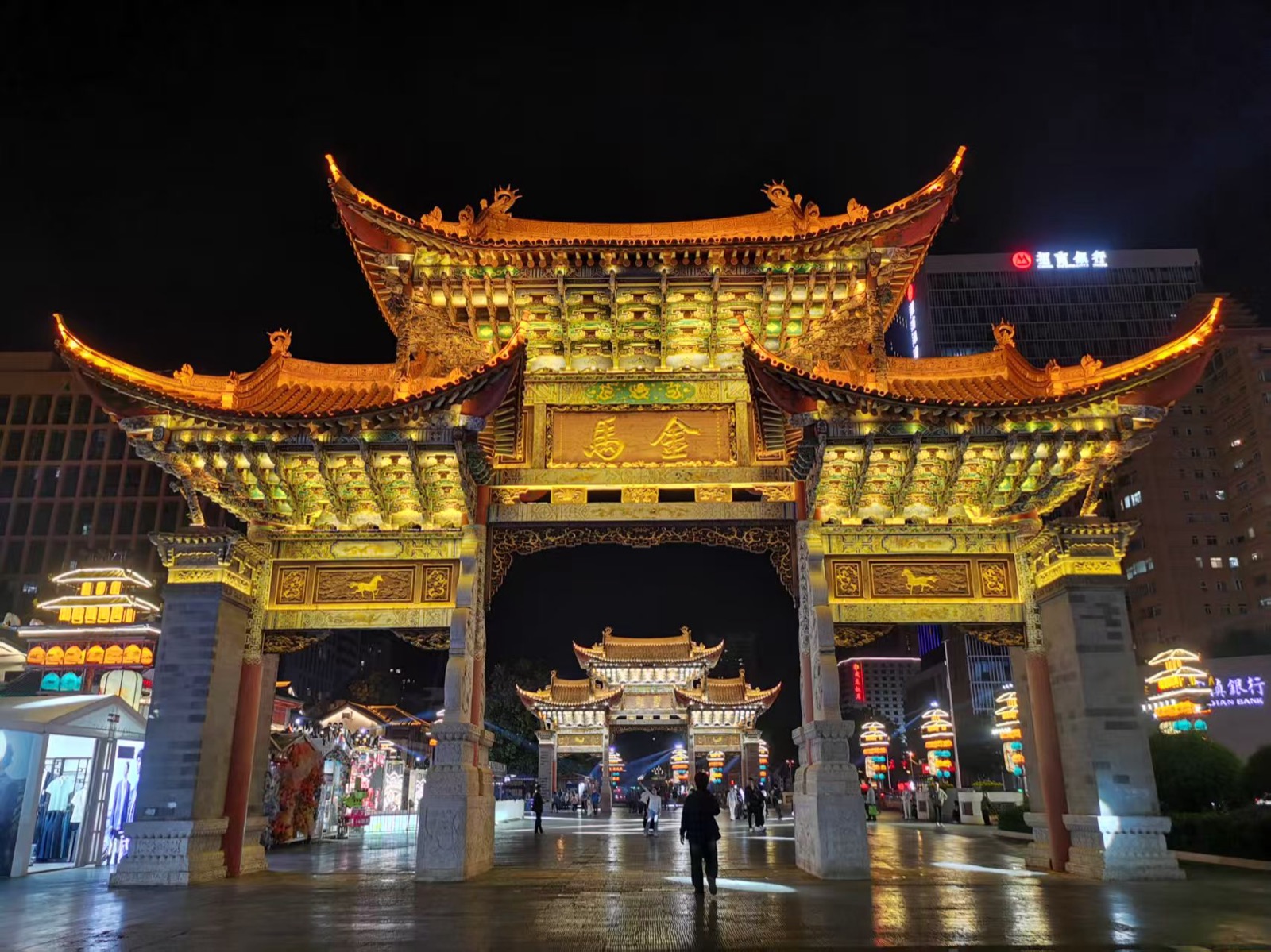 夜晚的昆明金马碧鸡坊，金色坊门飞檐翘角、灯火璀璨，远处城市楼宇与霓虹相映