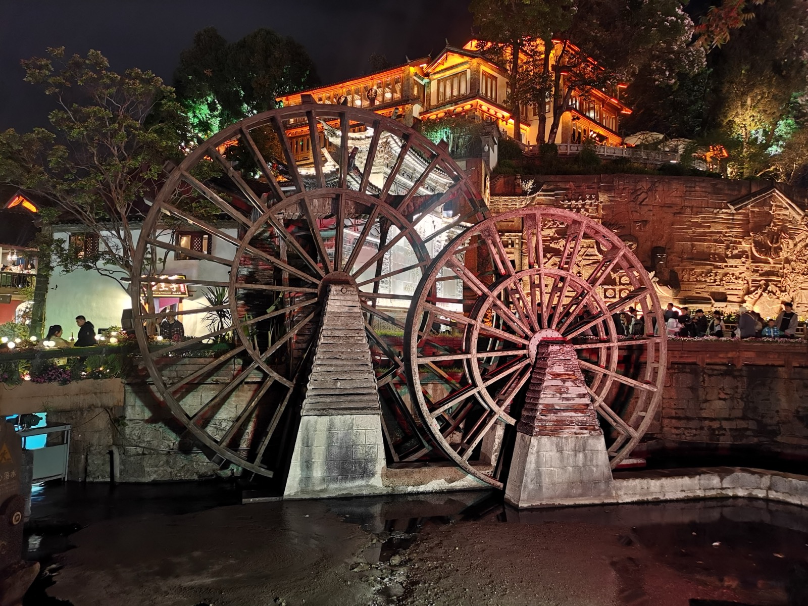 丽江古城入口处两座巨大的木质水车在夜色中亮着暖光，背后是灯火通明的古建筑