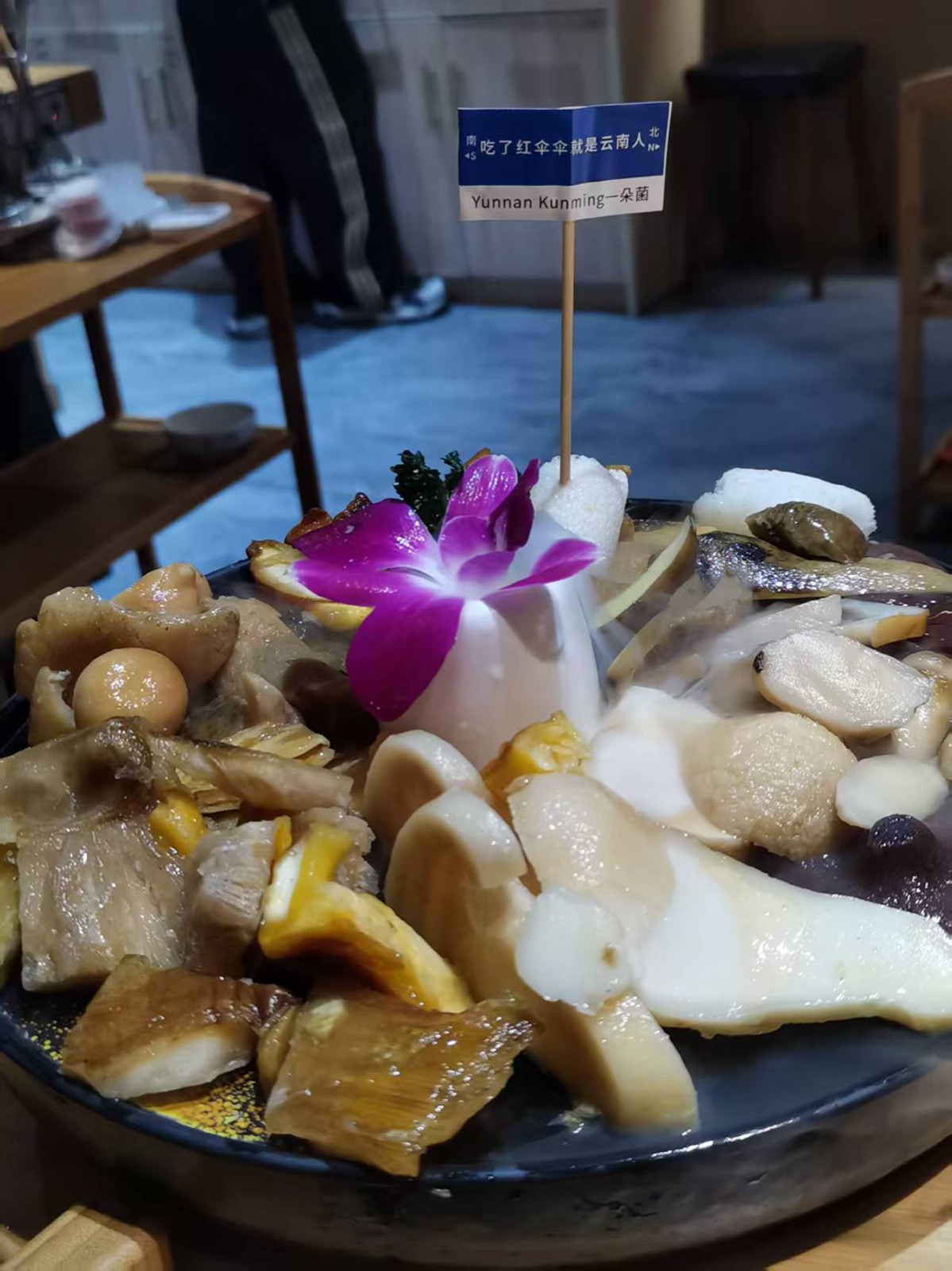 一大盘各色野生菌摆成拼盘，中央插着一朵紫色兰花和一面写着「吃了红伞伞就是云南人」的小旗