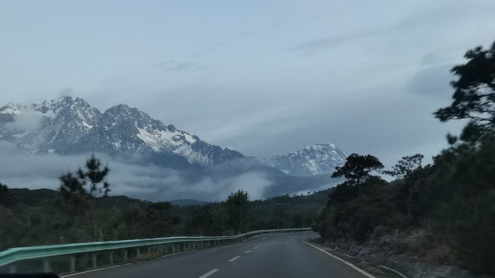 清晨公路尽头的玉龙雪山，雪峰被云雾半掩，山下是松林与蜿蜒山路